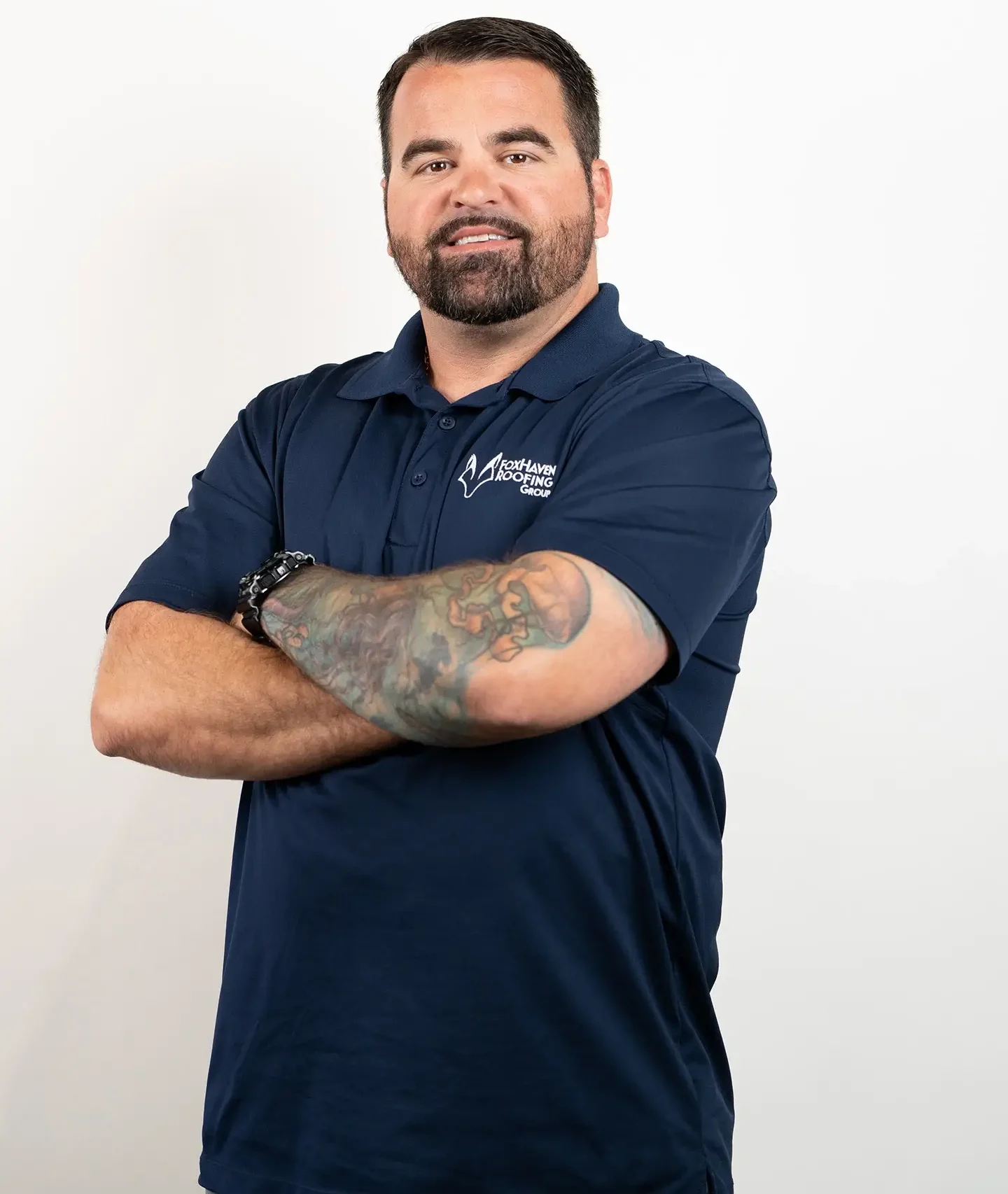 A man with dark hair and a beard, wearing a navy blue polo shirt with a logo, stands with his arms crossed. He has a tattoo sleeve on his left arm and is smiling against a plain white background.