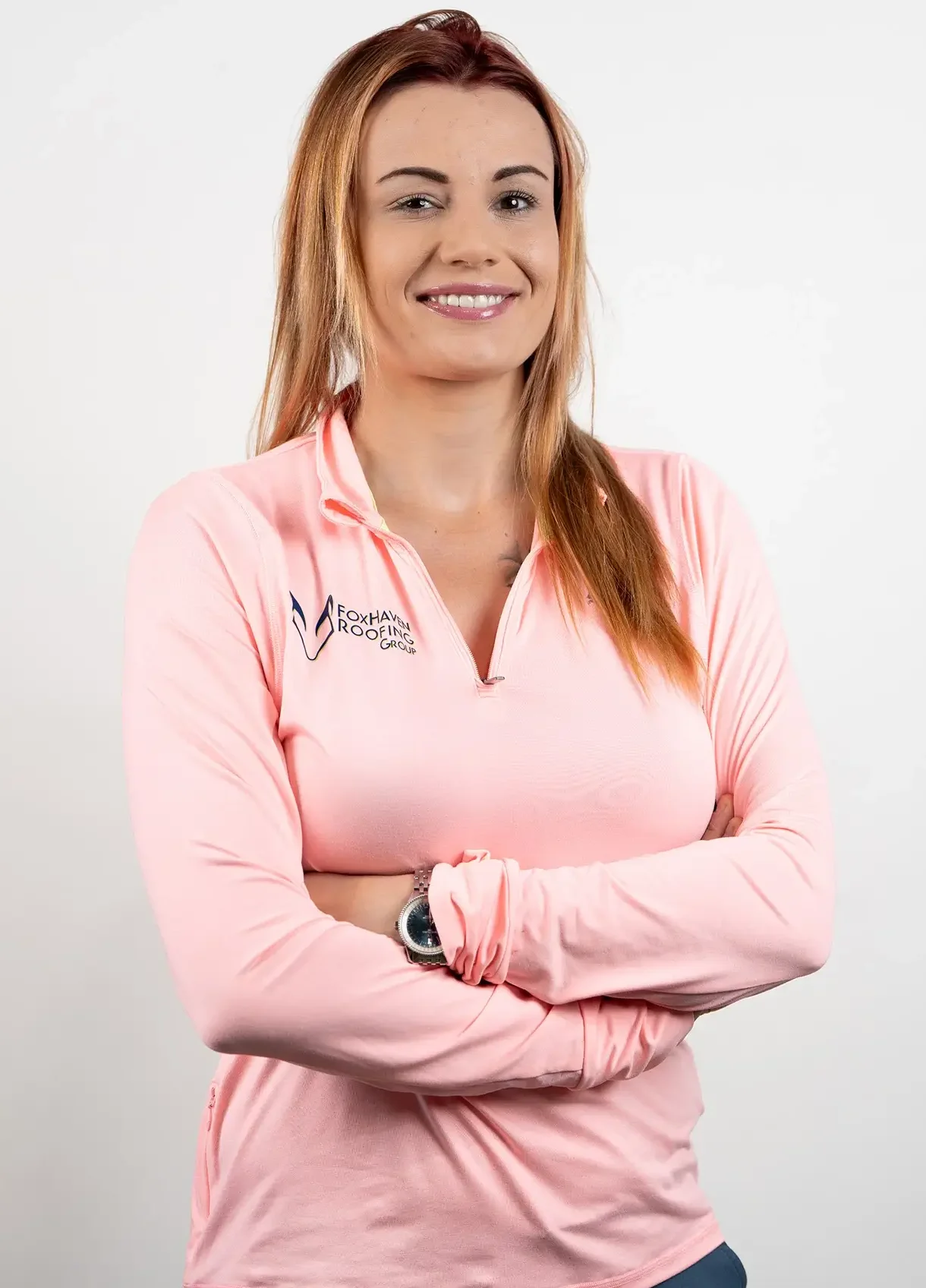 A woman with long, light brown hair smiles with her arms crossed. She is wearing a light pink long-sleeve shirt with the