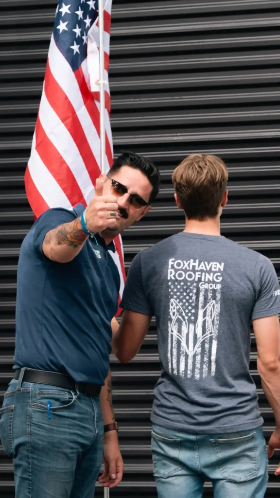 A man wearing sunglasses points toward the camera while holding an American flag. Another man stands beside him, back facing the camera, wearing a FoxHaven Roofing Group T-shirt. Both stand in front of a dark metal wall.