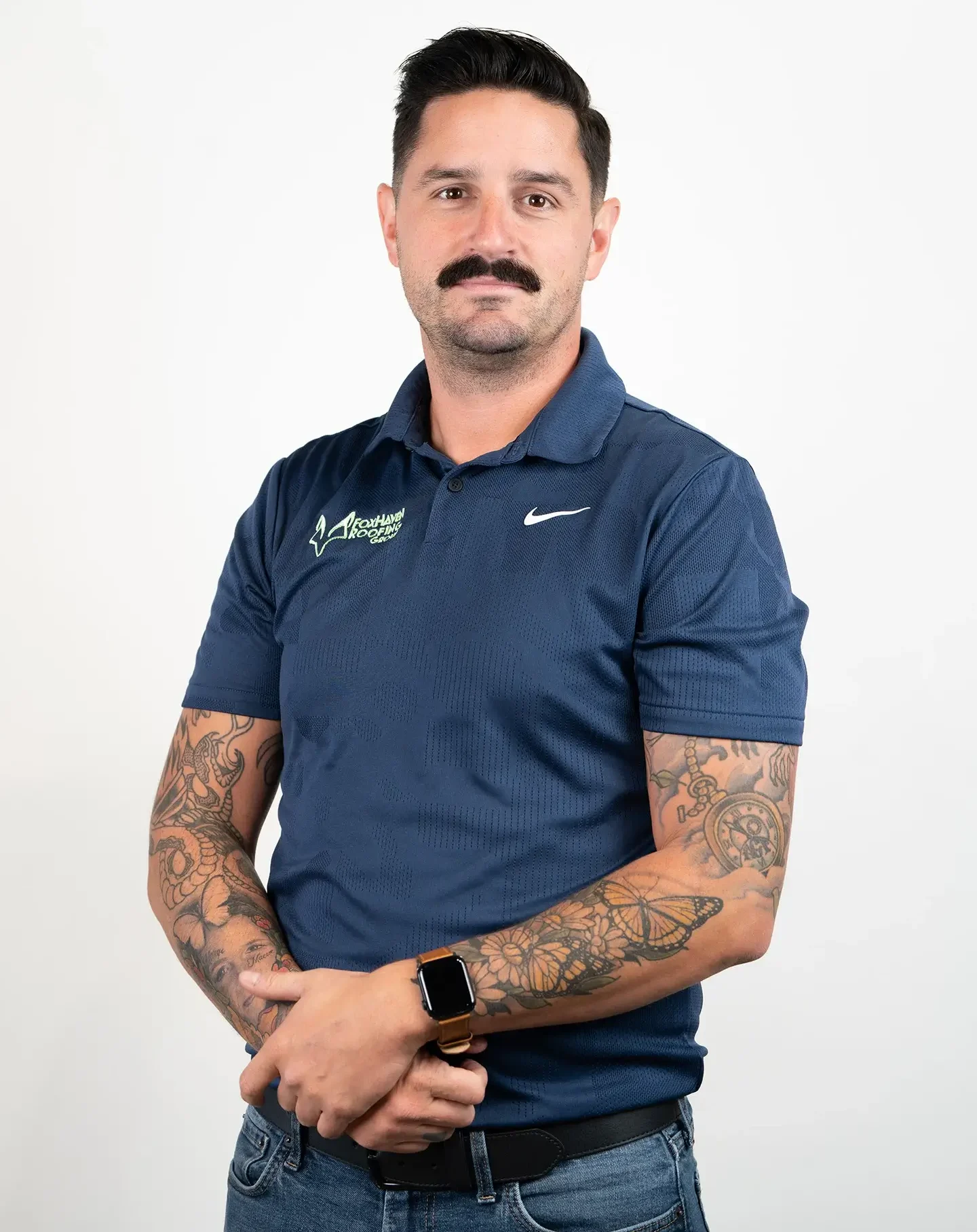 A man with dark hair and a mustache, wearing a navy blue polo shirt and jeans, stands with his arms crossed. He has visible tattoos on his forearms and wears a smartwatch. The background is plain white.