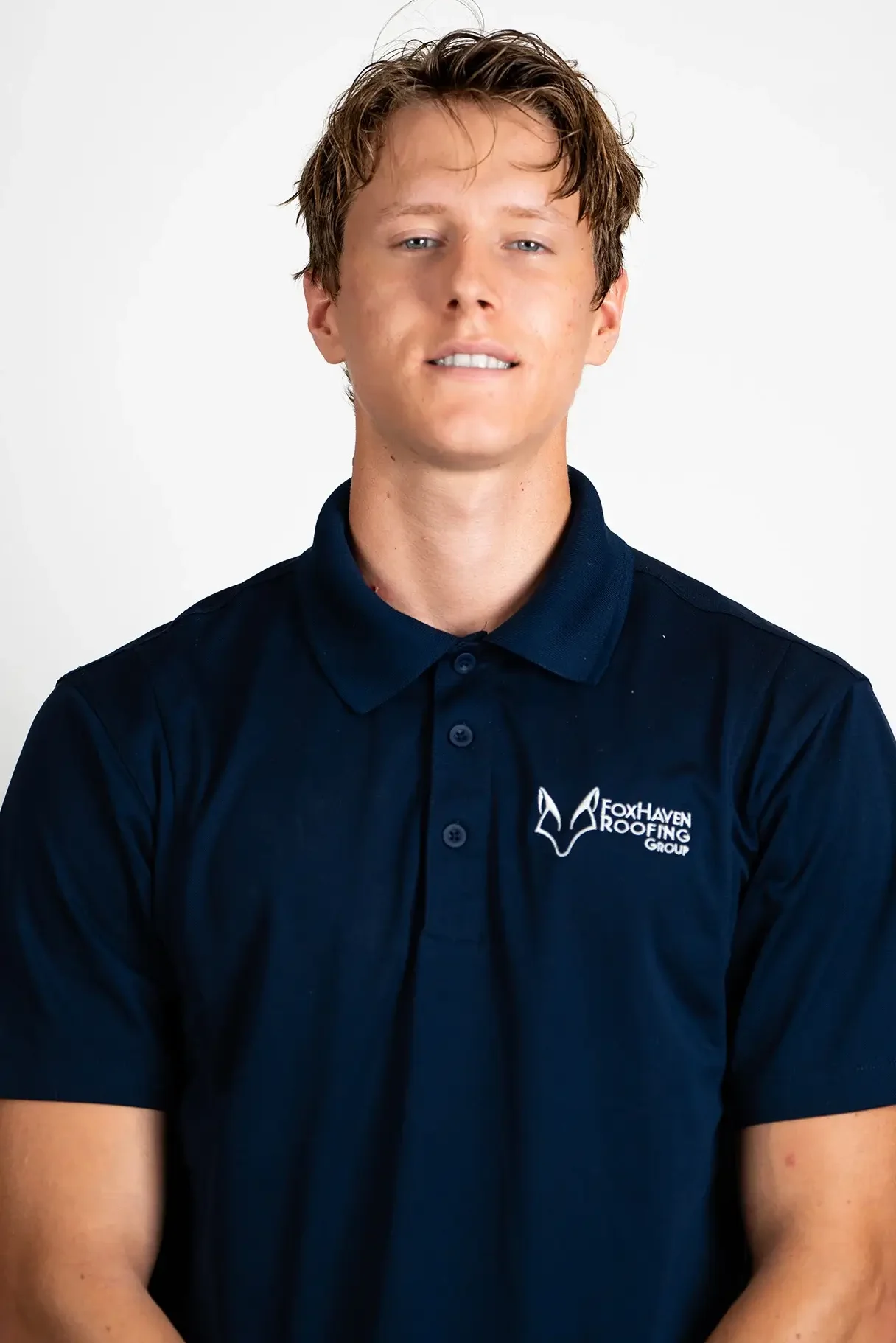 A young man with short, light brown hair smiles slightly while wearing a navy blue polo shirt with a white