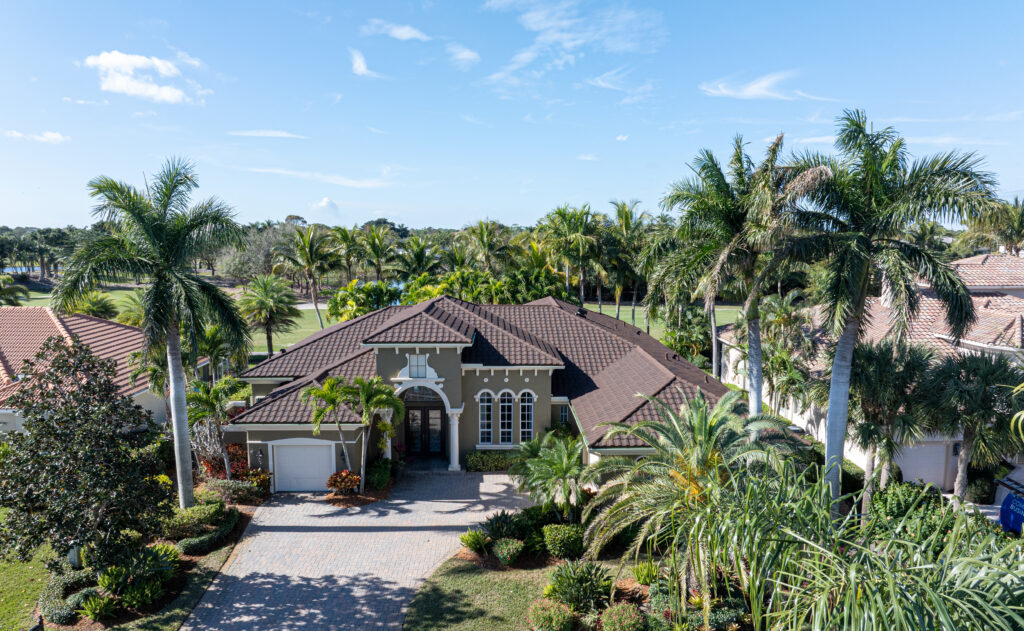 Aerial view of a large, single-story house with a brown tile roof, surrounded by palm trees and lush greenery, under a clear blue sky. The house has a paved driveway and manicured landscaping.