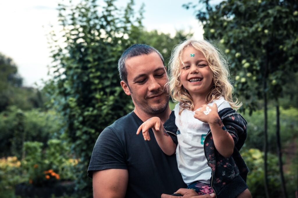 A man is holding a smiling young girl with blonde hair outdoors in a lush, green garden near metal roofing in Jupiter Farms. The girl is laughing, with stickers on her face and hand, and both appear happy and relaxed.
