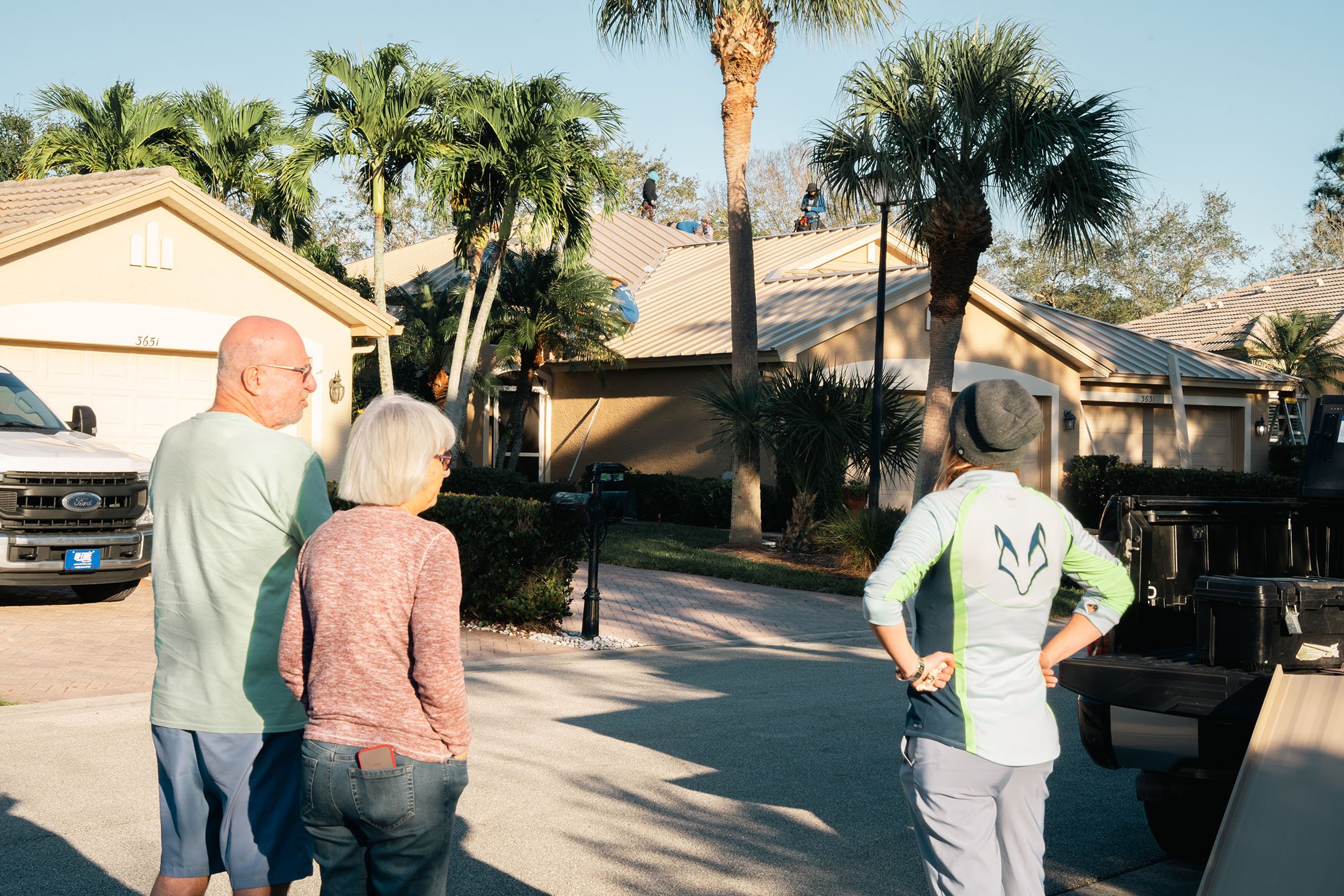Three adults stand on a residential street, looking toward workers installing solar panels on the roof of a house surrounded by palm trees. The scene is sunny, and houses with tan roofs line the street.