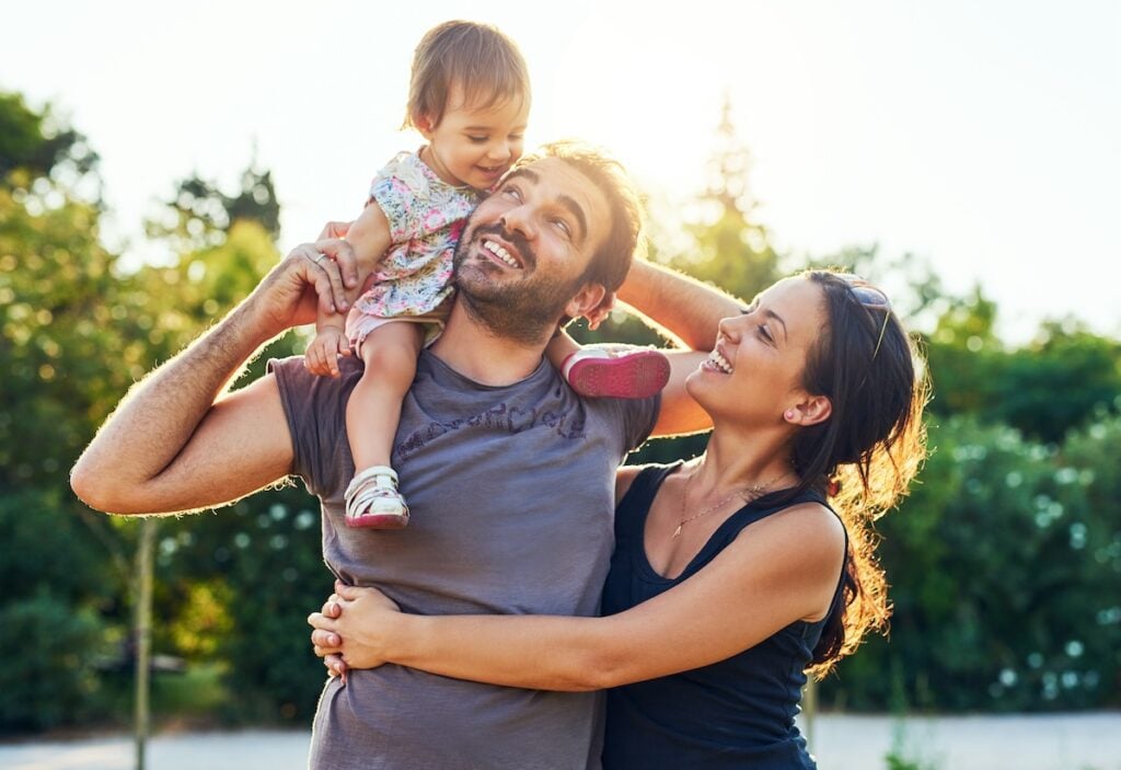 A smiling family stands outdoors in sunlight; an adult holds a laughing toddler on their shoulders while another hugs them, all enjoying the moment together. Trees and sunlight—plus glimpses of durable metal roofing Palm City homes—are in the background.