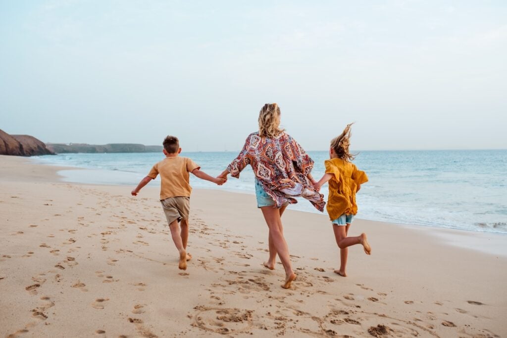 A woman and two children, all barefoot, run along a sandy beach near the ocean in Palm Beach Garden, holding hands. The sky is clear and calm waves are in the background. The group is seen from behind.