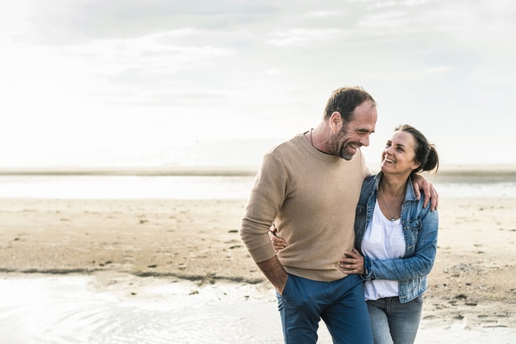 A smiling couple walks arm in arm along a sandy beach near Tequesta. The man wears a tan sweater and blue pants, and the woman wears a denim jacket and white top. They are looking at each other happily with the ocean—and roofing Tequesta—in the background.