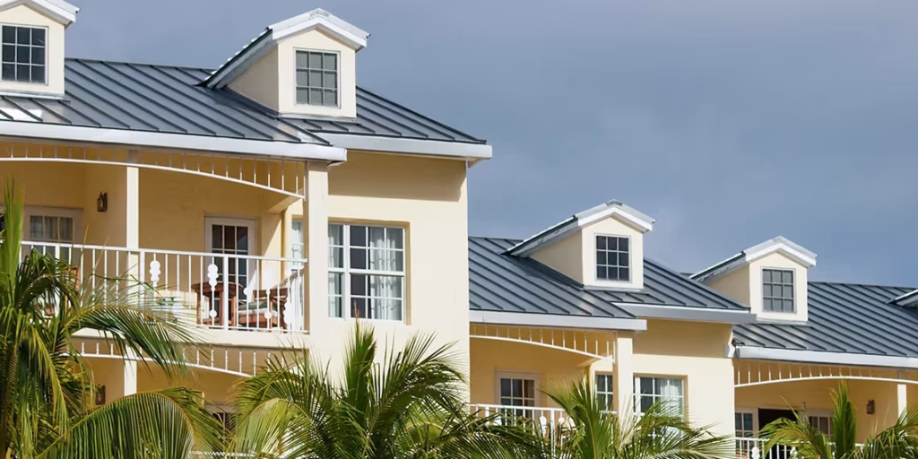 A row of yellow houses with white trim and metal roofs, featuring upper balconies and dormer windows, partially obscured by palm trees under a cloudy sky—a charming example of metal roofing Stuart style.