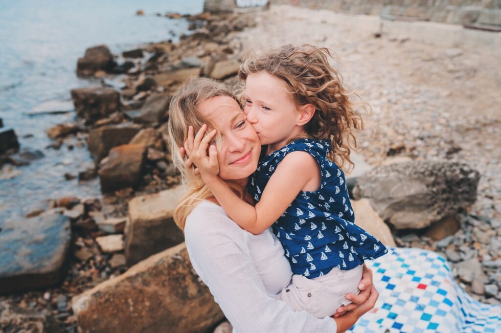 A young girl with curly hair kisses a smiling woman on the cheek while sitting on her lap at a rocky beach near Stuart. The woman gently holds the girl, and they both appear happy and affectionate under the clear sky, not far from local metal roofing in Stuart.