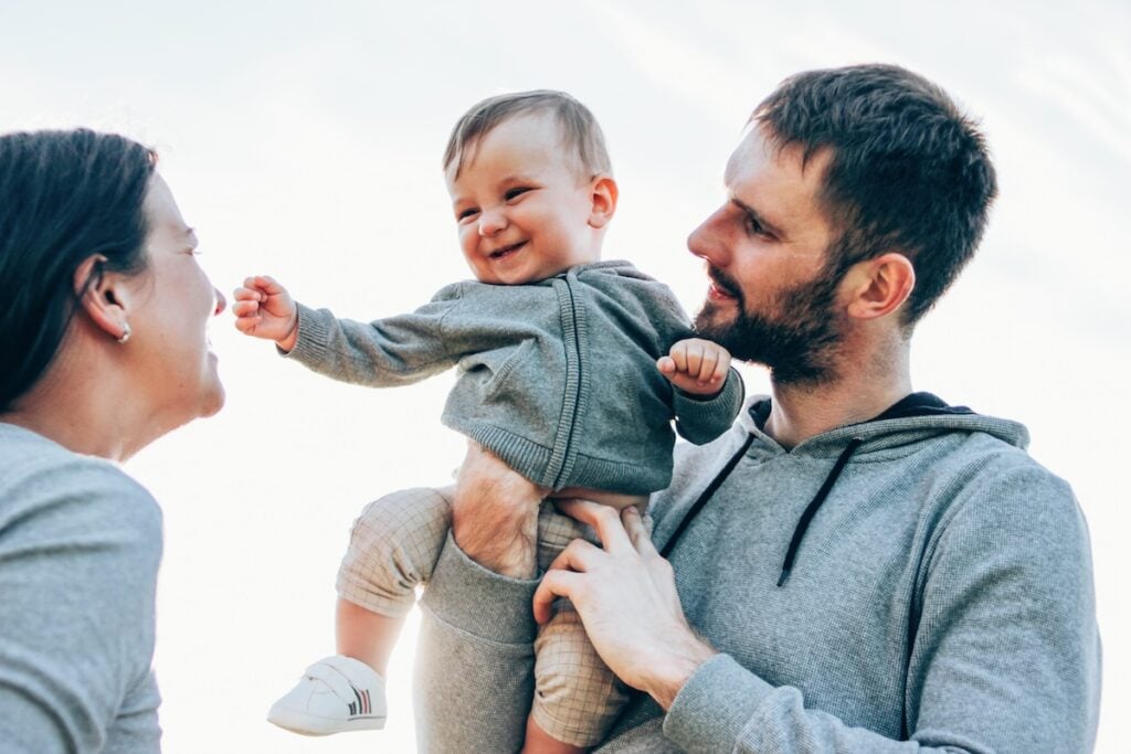 A smiling baby in gray clothes is held up between two adults, also wearing gray, outdoors under a clear sky—perfect weather for roofing Juno Beach. The baby laughs at one adult while the other gently supports the child.