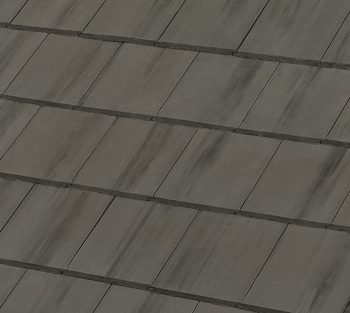 A close-up view of overlapping dark gray roof tiles arranged in a neat, diagonal pattern. The tiles have slight variations in shading, giving them a textured and weathered appearance.
