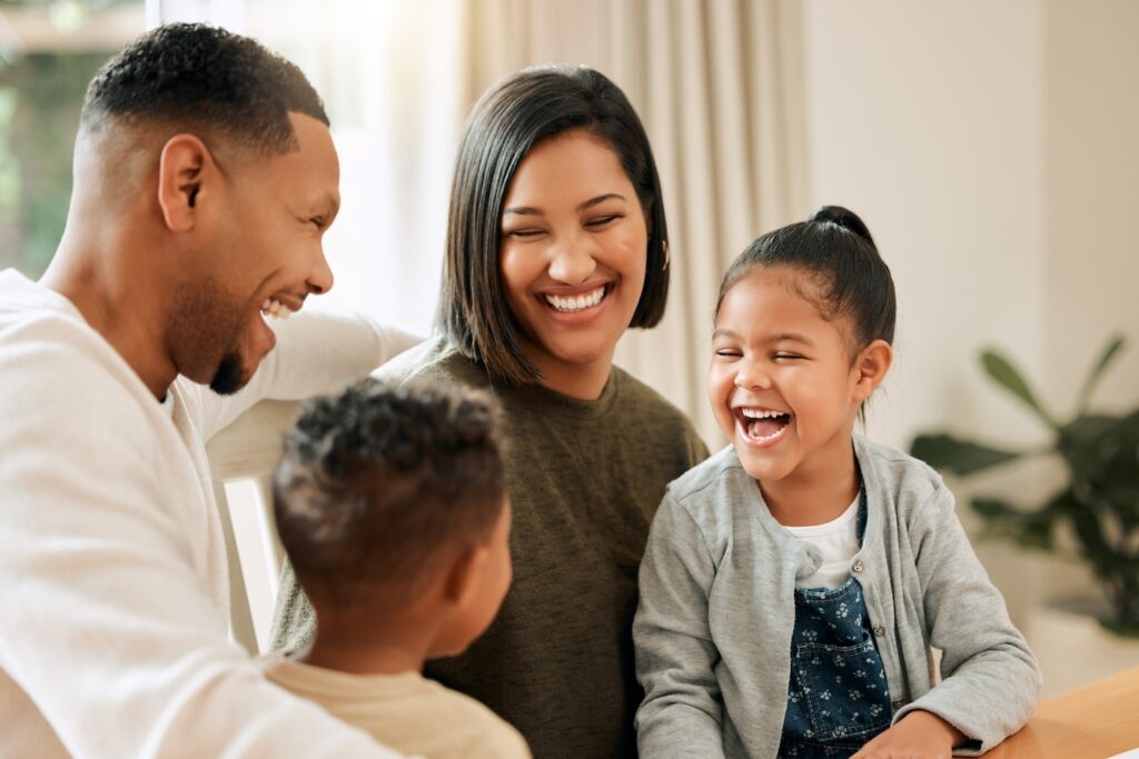 A happy family of four sits together indoors, smiling and laughing. The two parents look lovingly at their young son and daughter as they all share a joyful moment in their bright, cozy home with quality roofing Jupiter Island professionals trust.