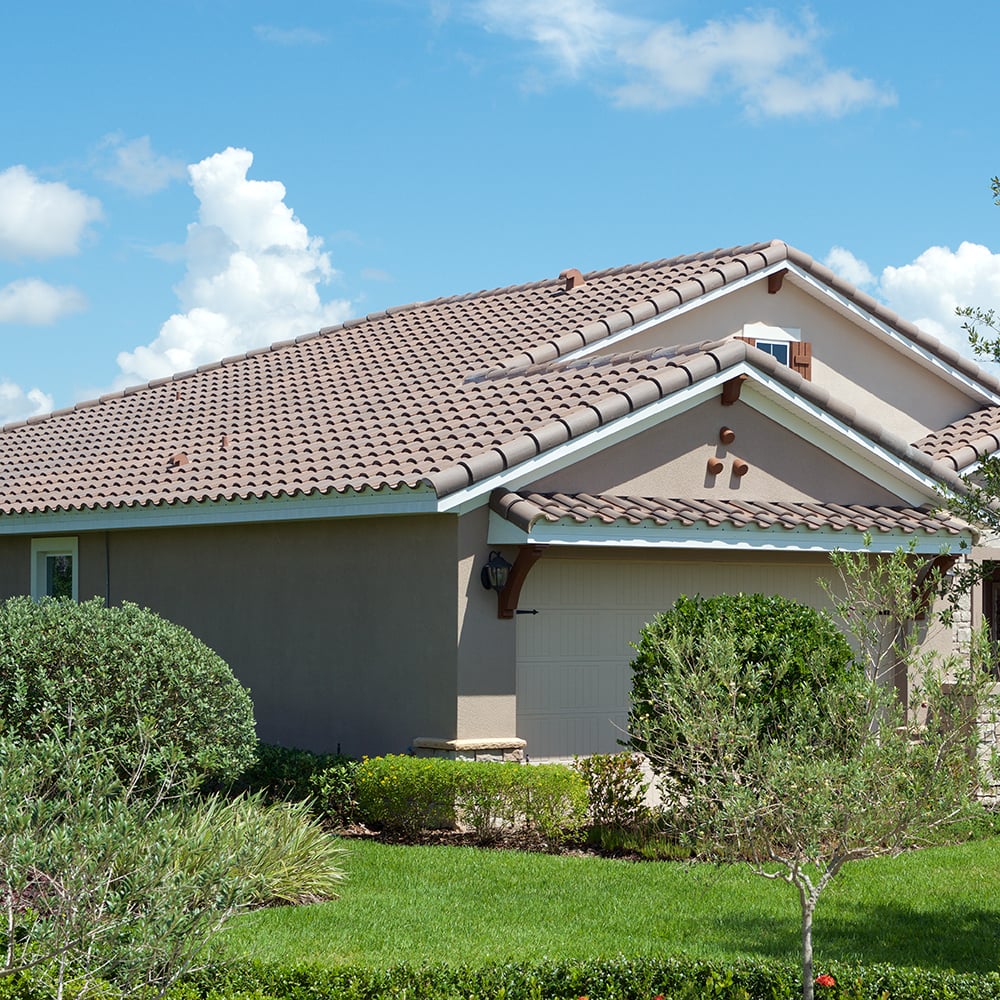 Single-story house with a beige exterior and brown tiled roof—expertly installed by roofing Jensen Beach—surrounded by a well-maintained green lawn, shrubs, and small trees under a bright blue sky with scattered clouds.