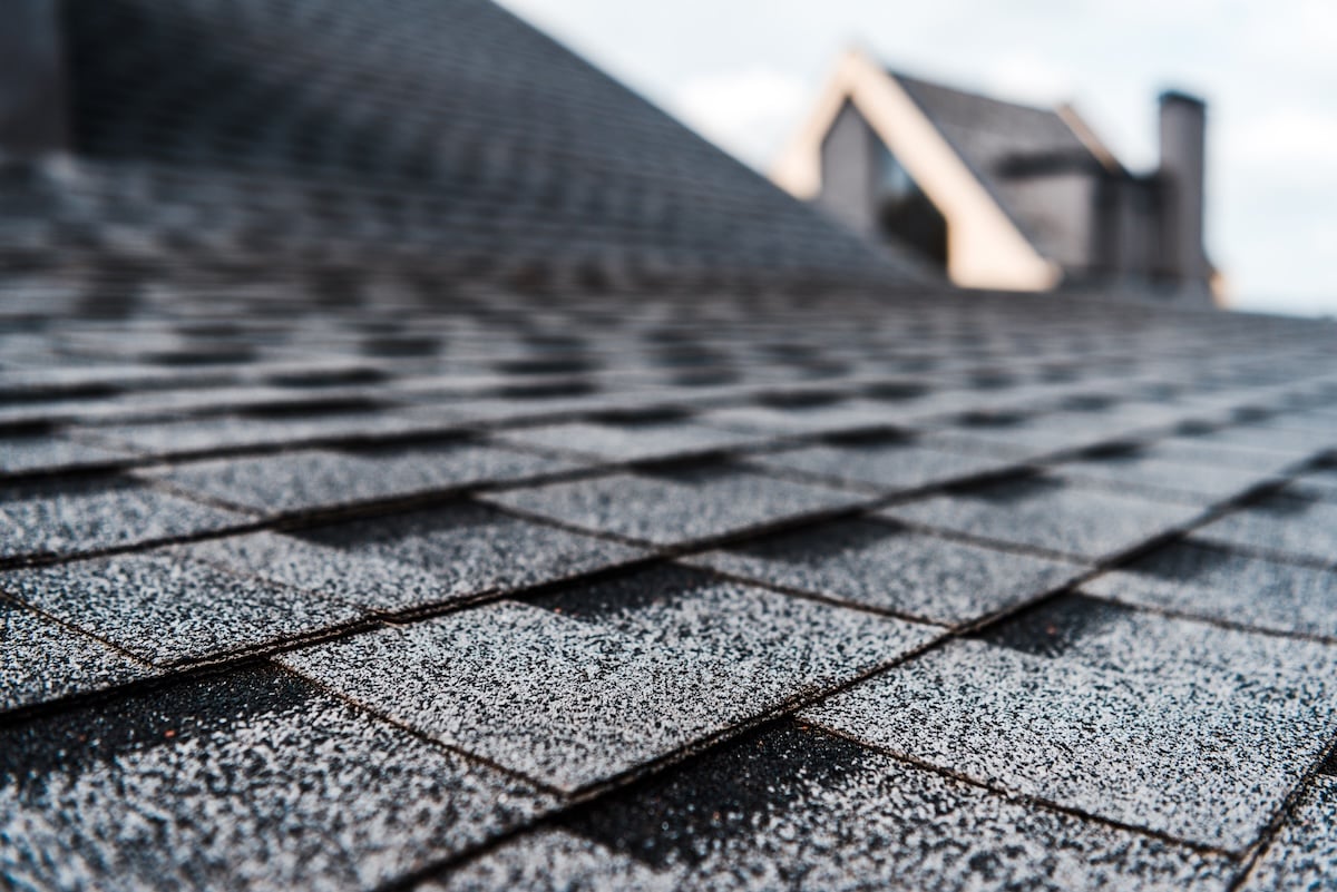 Close-up view of gray shingles on a sloped roof, with a blurry background showing the upper part of a house