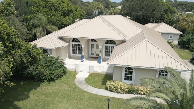 Aerial view of a light-colored, single-story house with a beige metal roof by roofing Jensen Beach, white trim, arched windows, and a landscaped yard surrounded by lush green trees and shrubs.