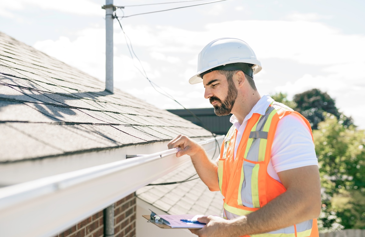 A worker performs roof inspection, checking a house’s gutter with a clipboard in hand while standing next to a sloped roof on a sunny day.