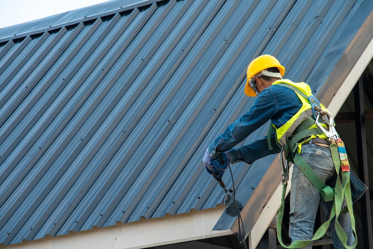 A construction worker performs metal roof repair, installing or inspecting the edge with tools and while standing on the building's structure.