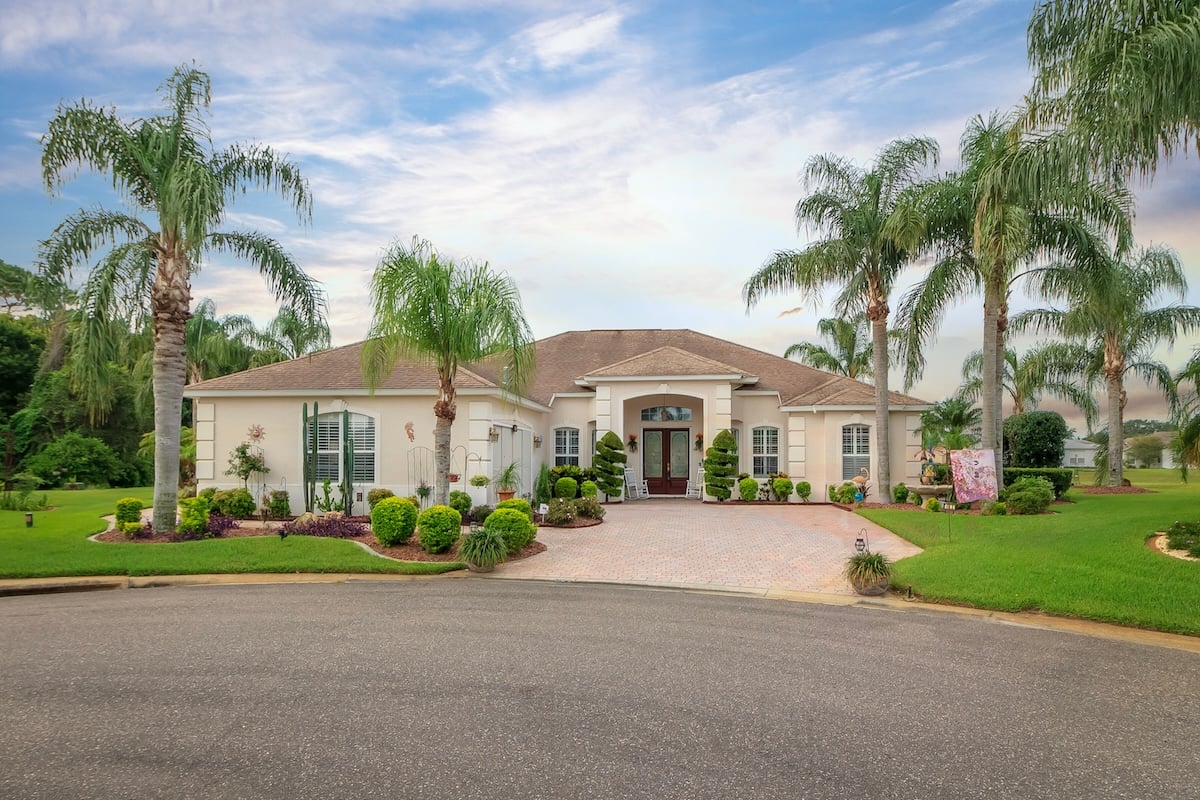 A single-story, beige house with roof shingles, a large driveway, manicured lawn, and palm trees in front