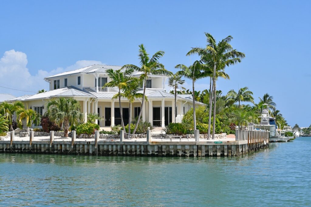 A large, white waterfront house with palm trees sits on a corner lot by a canal