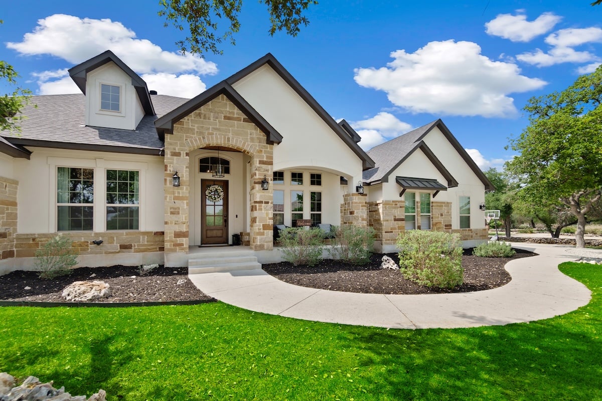 A modern suburban house with stone and cream siding, arched windows, and a dark gable roof. Manicured lawn and a curved walkway lead to the door, surrounded by shrubs and trees.