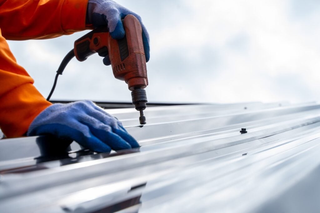 A person wearing blue gloves and an orange long-sleeve shirt uses a power drill to fasten screws into a sheet of metal roofing outdoors