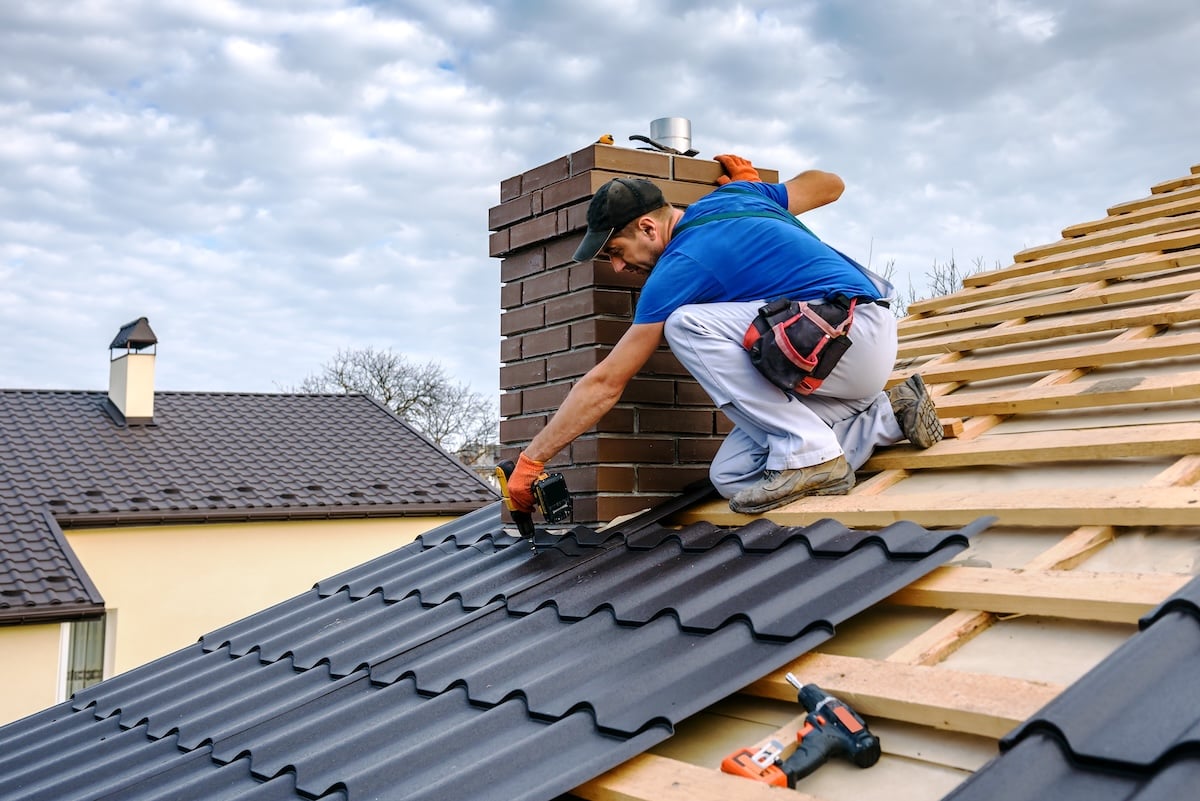 A worker installs black metal roofing sheets near a brick chimney on a sloped roof under a partly cloudy sky.