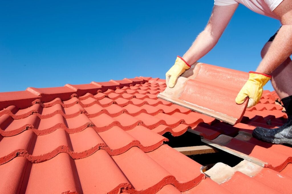 Roof repair, worker with yellow gloves replacing red tiles or shingles on house with blue sky as background