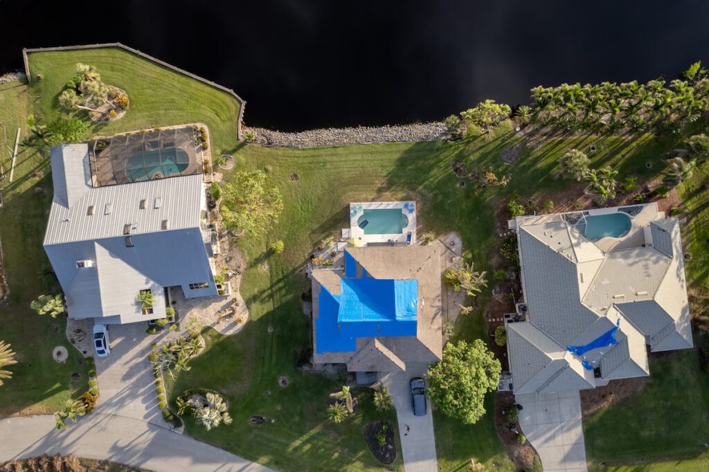 Aerial view of three houses with swimming pools in fenced backyards; two homes display blue tarps, indicating recent storm damage roof repair