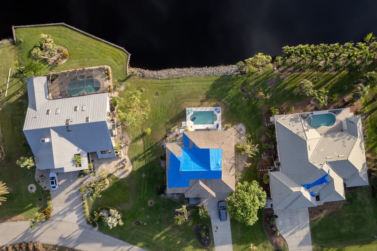 Aerial view of three houses with swimming pools in fenced backyards; two homes display blue tarps, indicating recent storm damage roof repair