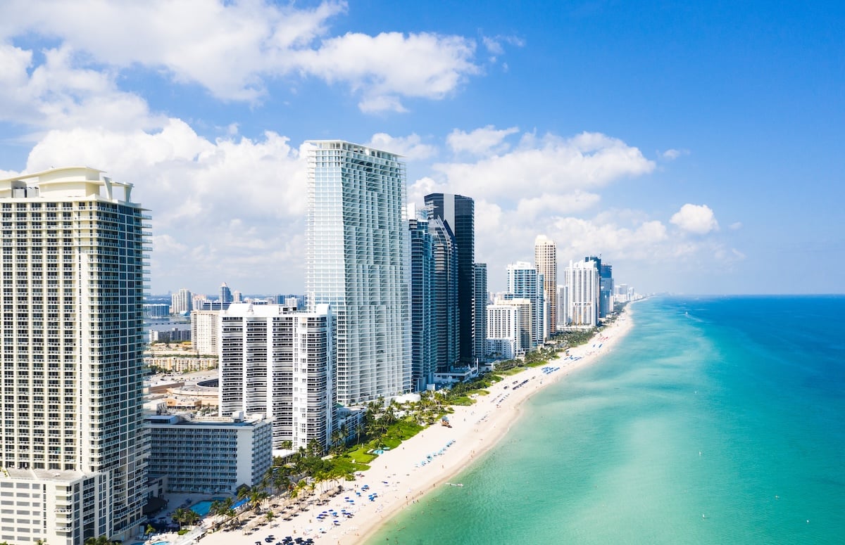 Aerial view of tall modern beachfront skyscrapers lining a white sandy beach with turquoise ocean waves