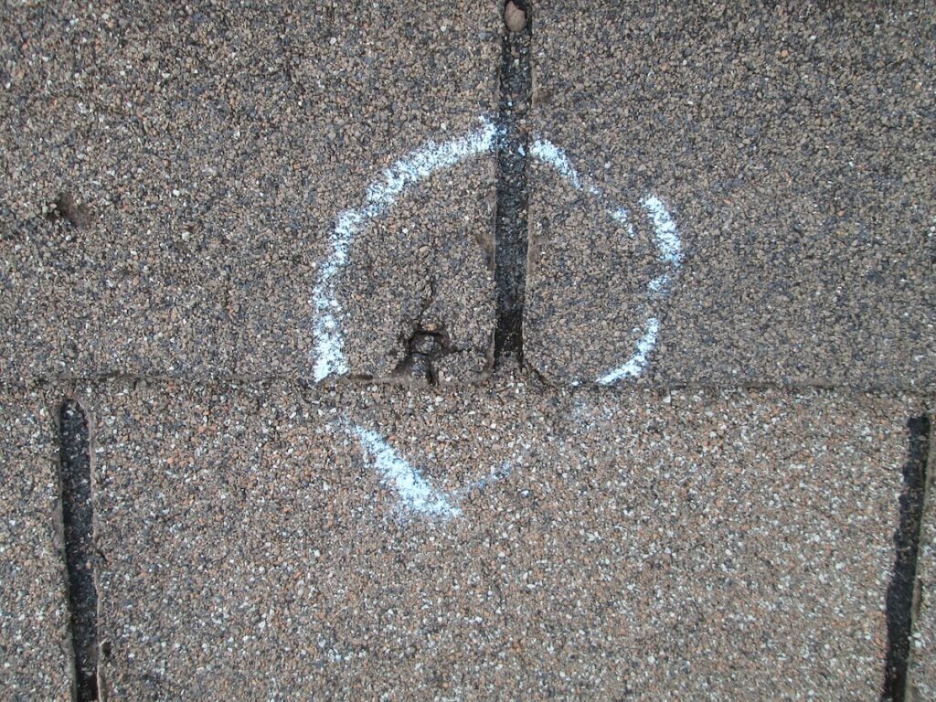 A section of asphalt roof shingles with a white chalk circle marks hail damage