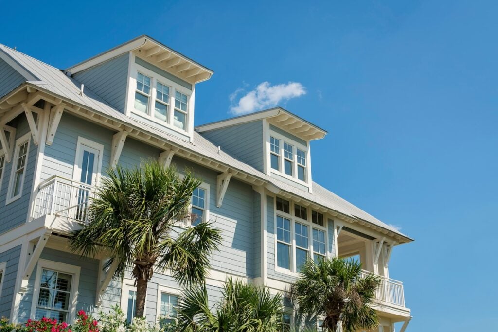 Light blue coastal-style house with white trim and large windows, featuring a balcony and surrounded by palm trees