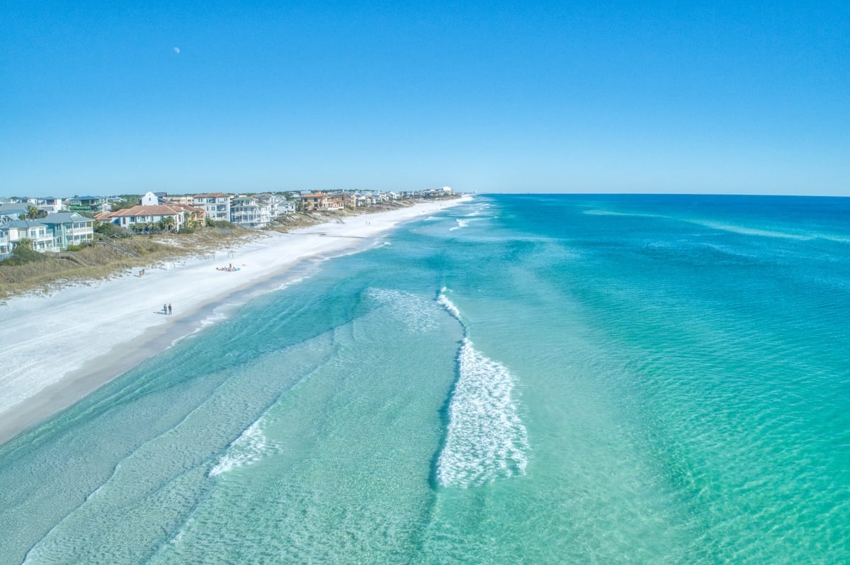 Aerial view of one of sandy shoreline with clear turquoise water, gentle waves, and houses along the coast under a bright blue sky.