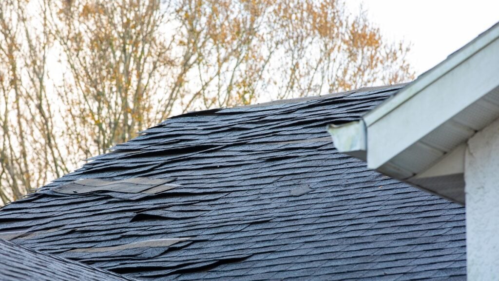 A close-up of a house roof with damaged, curling, and missing asphalt shingles, suggesting the need for storm damage roof repair