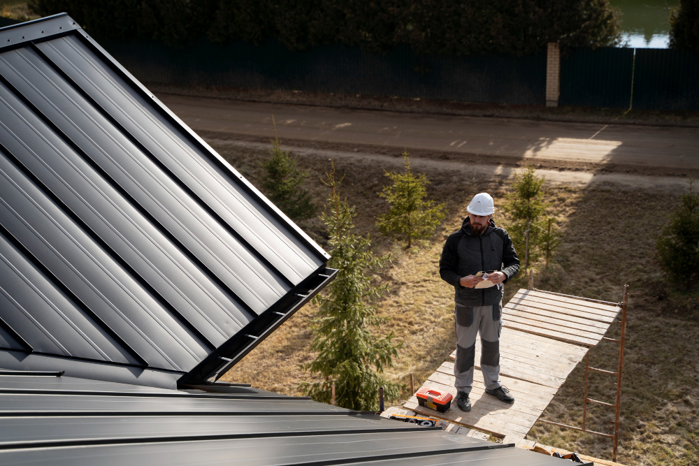 A construction worker wearing a white helmet stands on scaffolding next to a black metal roof, holding a clipboard and looking up, with trees and a road in the background.