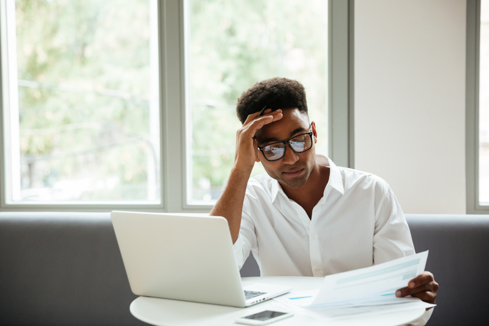 A man wearing glasses and a white shirt sits at a table with a laptop and smartphone, holding documents and looking stressed, with his hand on his forehead. Large windows are in the background.
