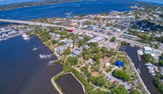 Aerial view of a coastal town with houses, roads, a marina, green spaces, and a bridge extending over blue water, with boats visible on the water and buildings lining the shoreline.
