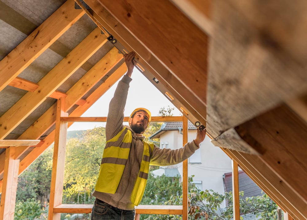 A construction worker in a yellow vest and hard hat uses a level tool to check the angle of a wooden beam inside a partially built house. Sunlight streams through the open frame.