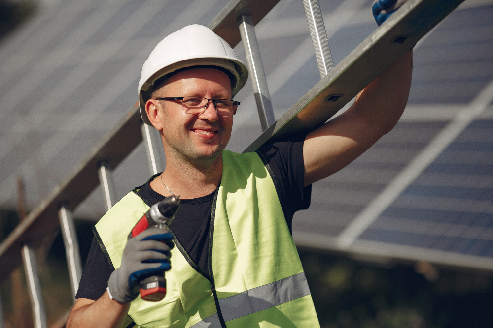 A smiling worker in a white hard hat and yellow safety vest carries a ladder on his shoulder and holds a power drill, with solar panels in the background.