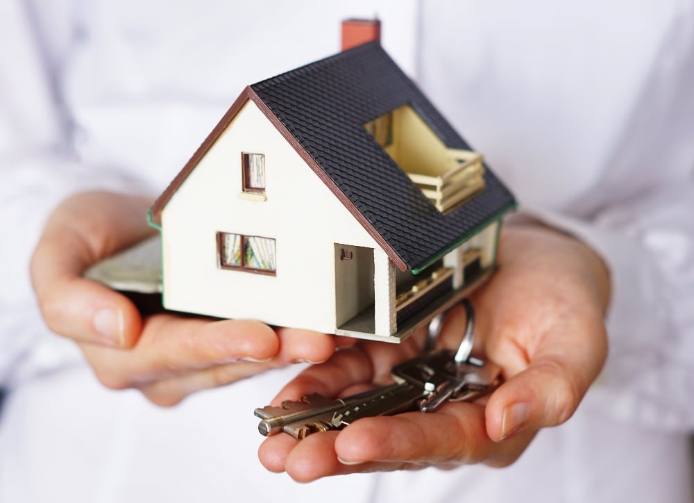 A person holding a small model house in one hand and a set of keys in the other, symbolizing real estate, home ownership, or buying a new home.