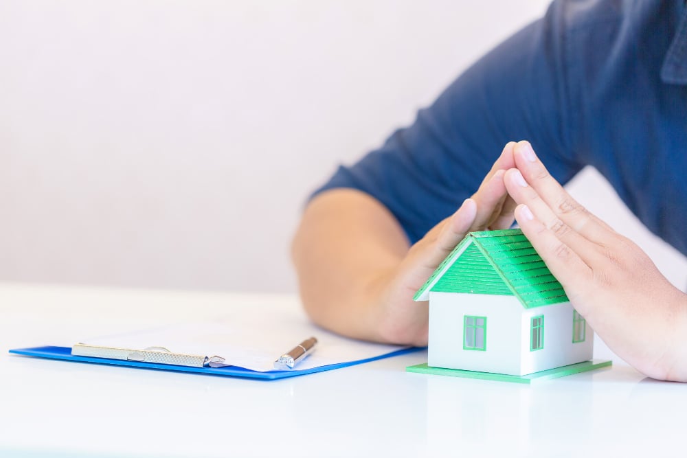 A person’s hands are cupped protectively over a small model house on a desk, next to a blue clipboard with papers and a pen.