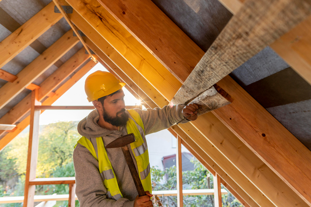 A construction worker in a yellow hard hat and reflective vest uses a hammer to secure a wooden board inside a house’s unfinished roof structure, with sunlight shining through the beams.