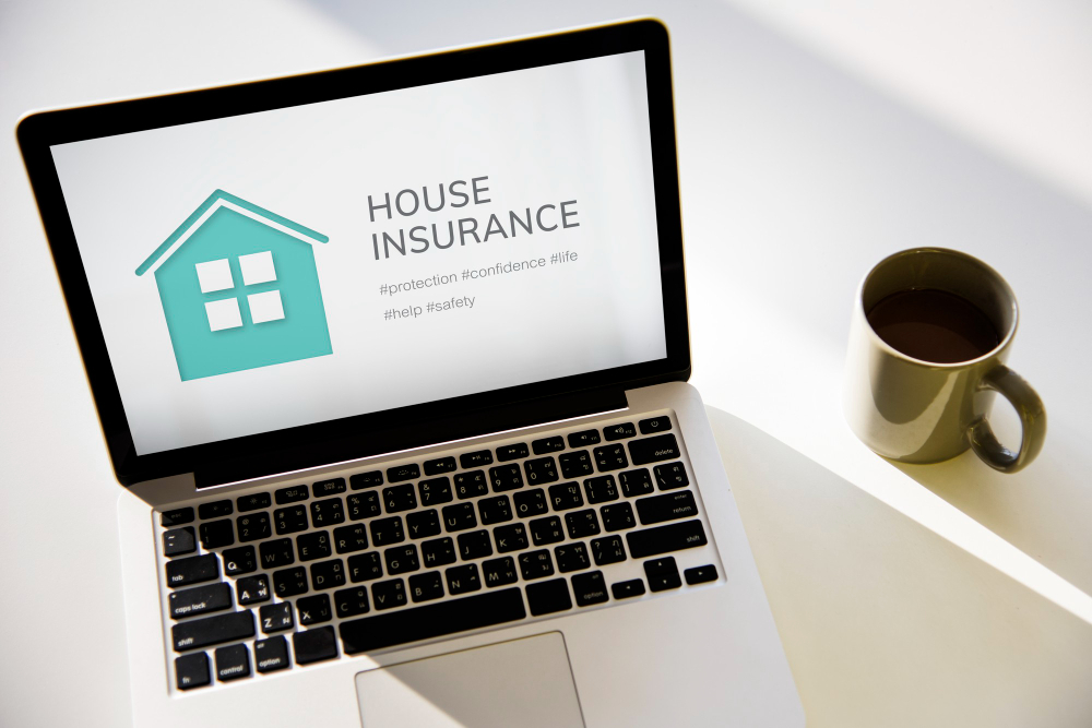 A laptop displaying a house insurance advertisement with a house icon and keywords. Beside the laptop is a cup of coffee on a white surface, bathed in natural light.