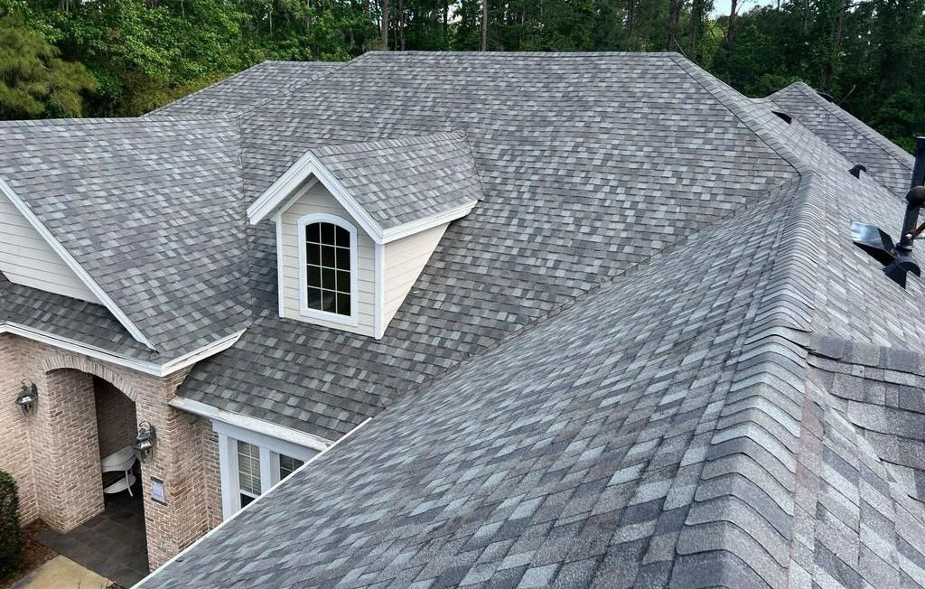 A large house with a complex gray shingle roof featuring multiple peaks, ridges, and a dormer window. The roof appears new and clean, surrounded by trees and greenery.
