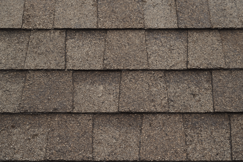 Close-up view of brown asphalt roof shingles arranged in horizontal overlapping rows, showing a rough, textured surface.