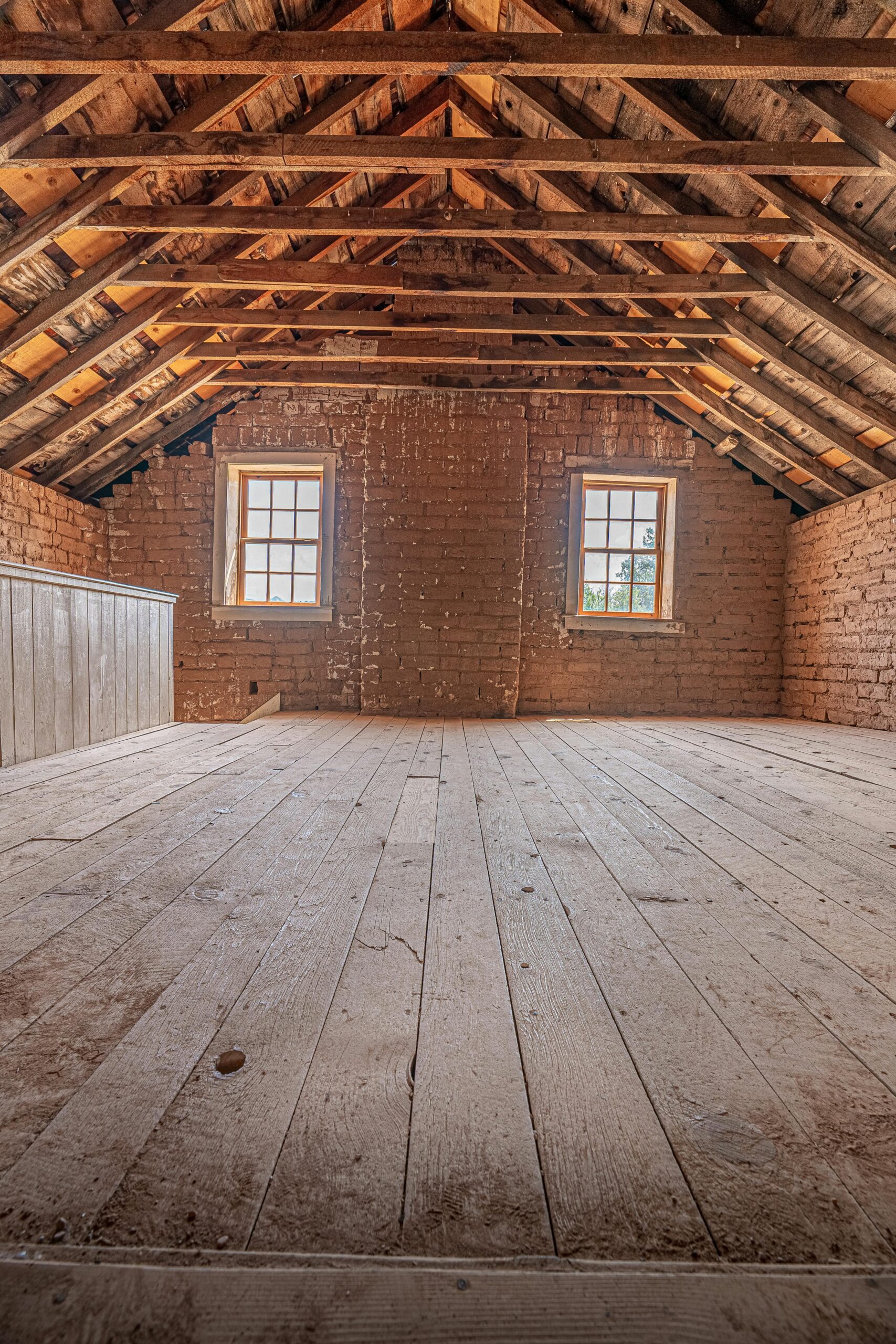 An empty attic with wooden floors, exposed beams, brick walls, and two windows letting in natural light. The space appears rustic and unfinished.