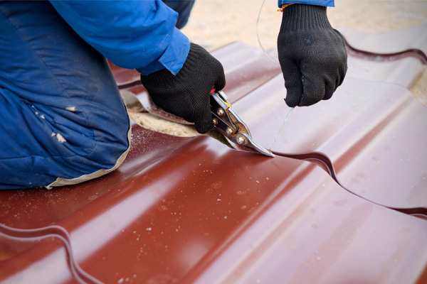 A person wearing black gloves and blue work clothes uses metal shears to cut a brown corrugated metal roofing sheet.