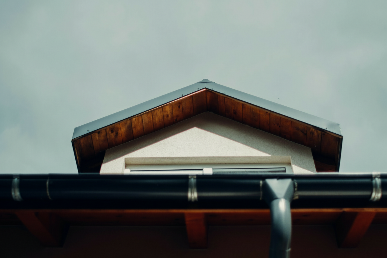 A close-up view of a house roof with a triangular gable, wooden eaves, and a metal gutter system, set against a cloudy sky.