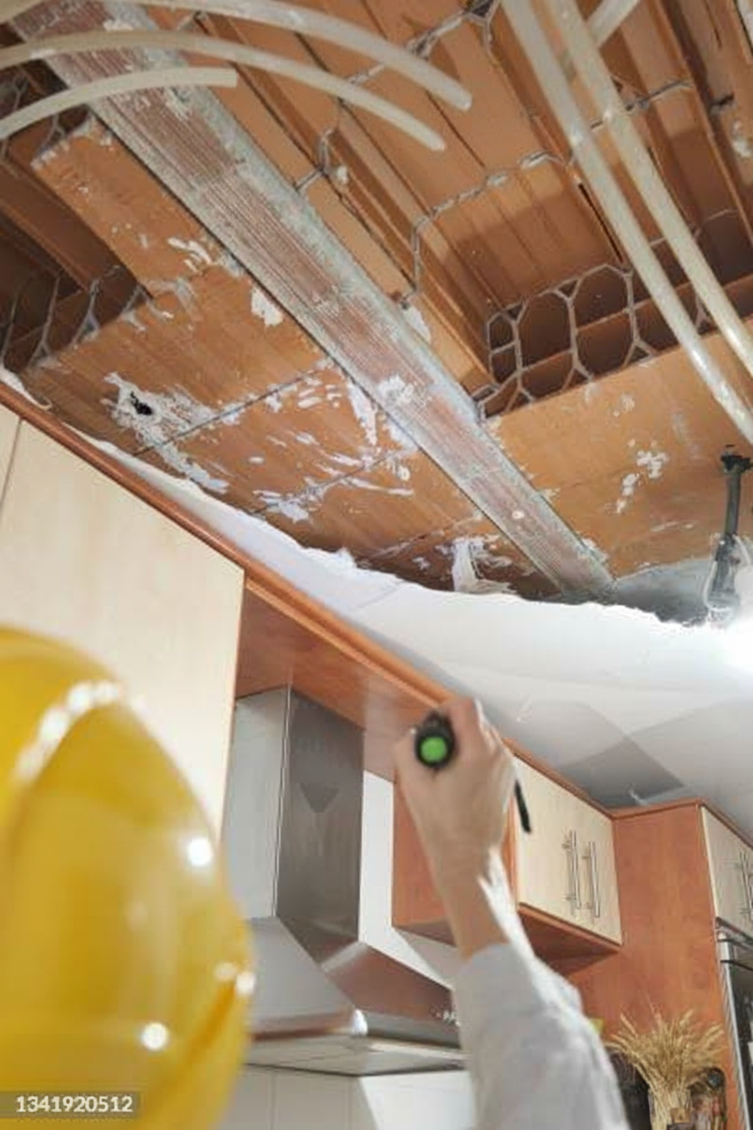 A person wearing a yellow hard hat inspects a kitchen ceiling, which shows significant water damage, mold, and exposed piping, using a flashlight.