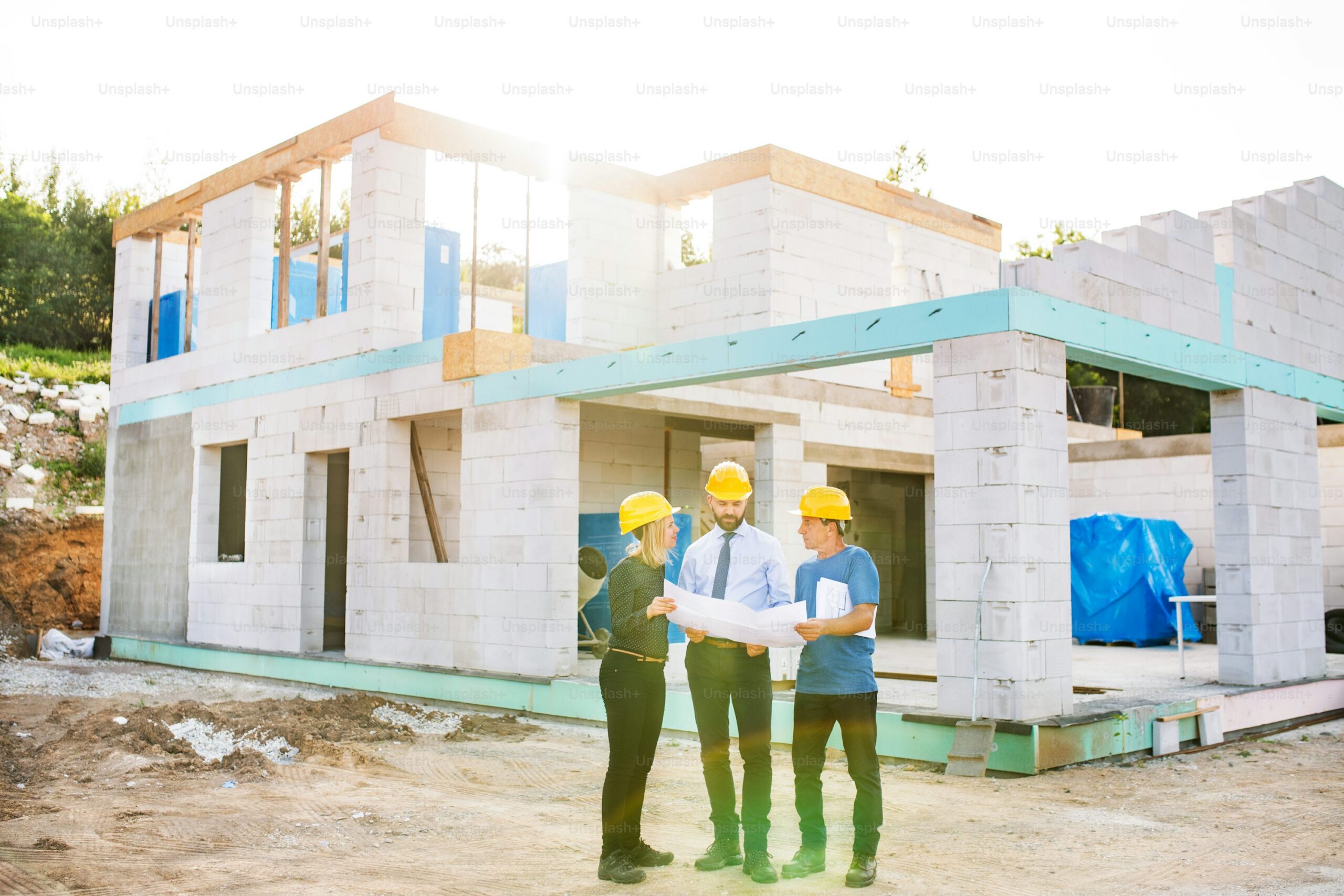 Three construction workers wearing yellow helmets stand in front of a partially built house, reviewing blueprints together at a construction site on a sunny day.